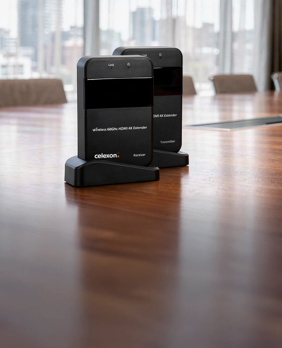 In the foreground, a celexon Expert Wireless HDMI Set WHD30M – 4K is placed on a large wooden table in a modern conference room, shown clearly and sharply. In the softly blurred background, two business professionals are conversing in front of floor-to-ceiling windows with a city view, while a presentation display is visible on the wall.
