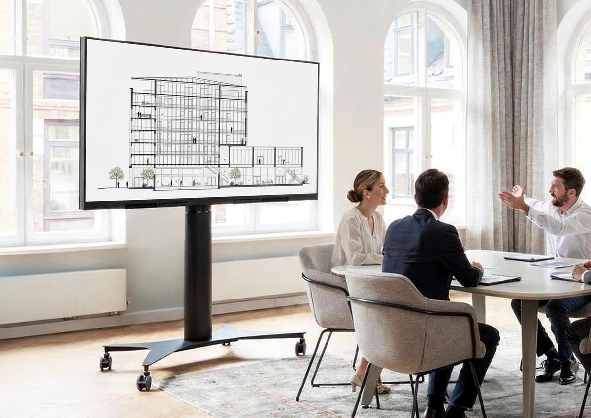 Four people are seated at a table in a bright, modern room, holding a meeting. On the left stands a celexon Expert electrically height-adjustable display trolley Adjust 42100M, showing an architectural drawing.