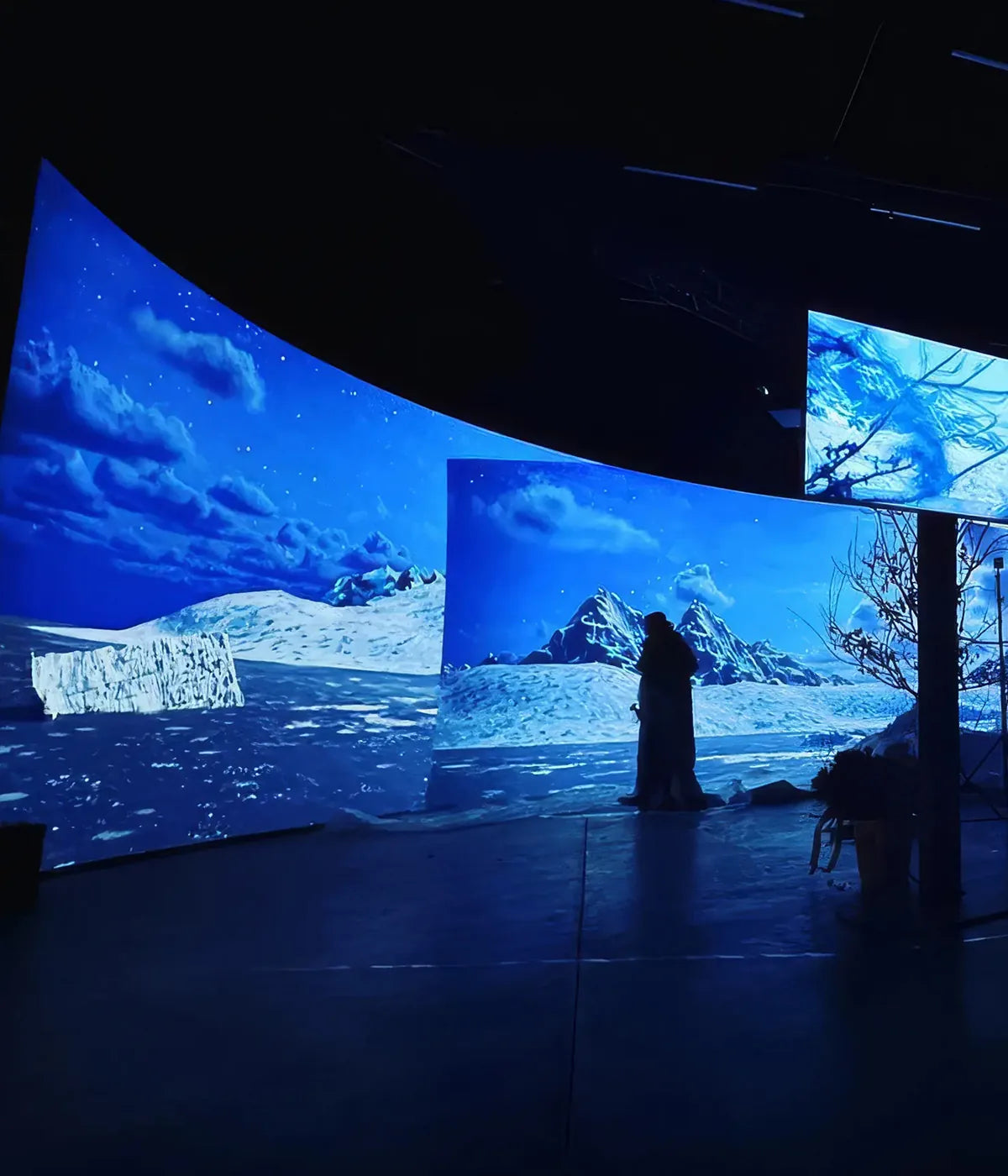 A large, dark studio with multiple curved projection screens displaying an icy polar landscape with mountains and sea; in front of them stand several silhouetted people and film equipment.