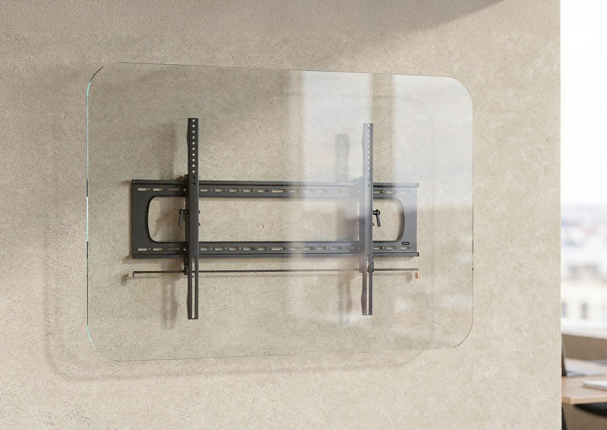The image shows the celexon TV/display wall mount Adjust/Fixed attached to a transparent glass panel. The bracket consists of a sturdy metal construction with horizontal and vertical rails for mounting a screen. The scene is set in a modern office or workspace with a neutral wall, large windows, and a desk in the background.