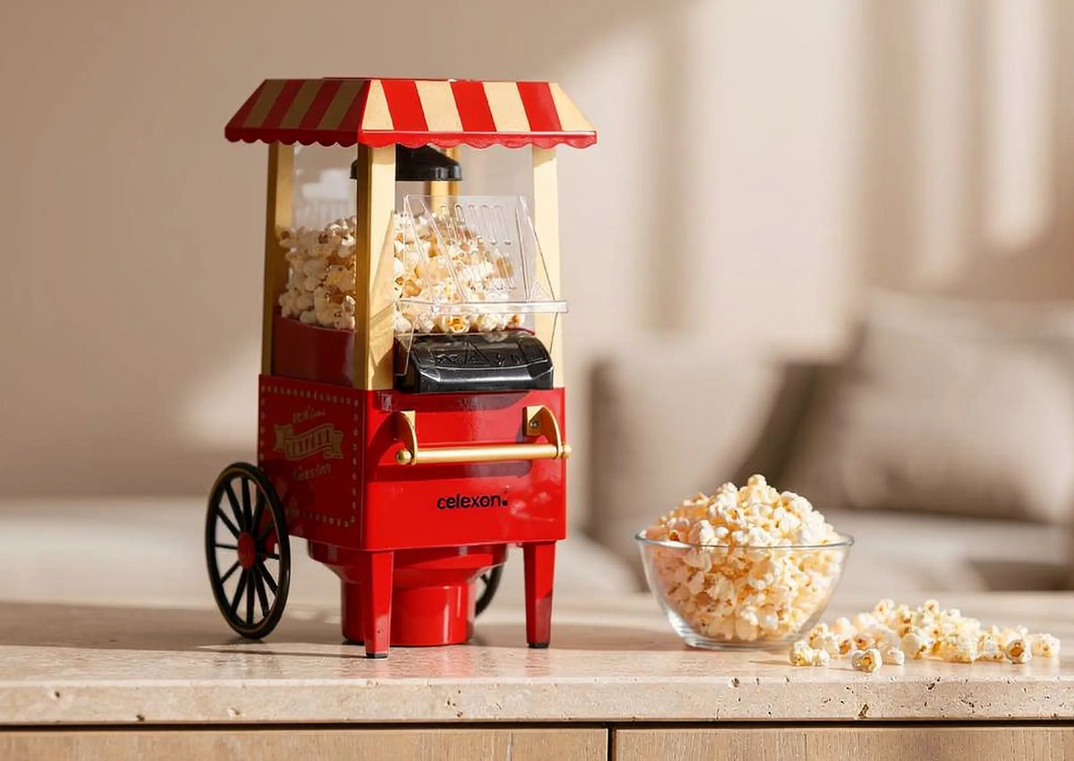 In the foreground stands a small, red celexon CP500 popcorn machine in the nostalgic style of a fairground cart with a red-and-white striped roof, filled with freshly popped popcorn. Beside it is a glass bowl of popcorn, with a few kernels casually scattered across the light wooden surface.