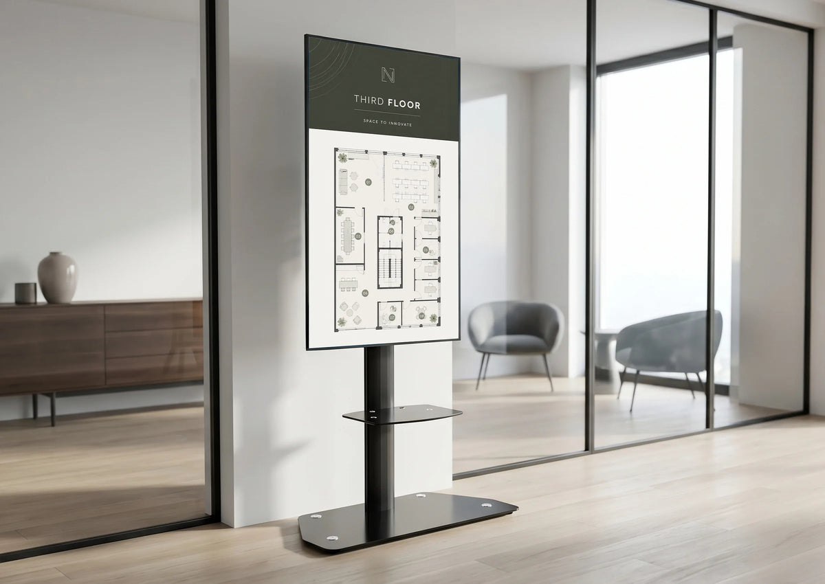 A modern, bright office with glass walls and minimalist furnishings; in the center stands a digital information display on a celexon Professional height-adjustable display trolley Adjust-3270MP Portrait, showing a building floor plan and the label “Third Floor.”