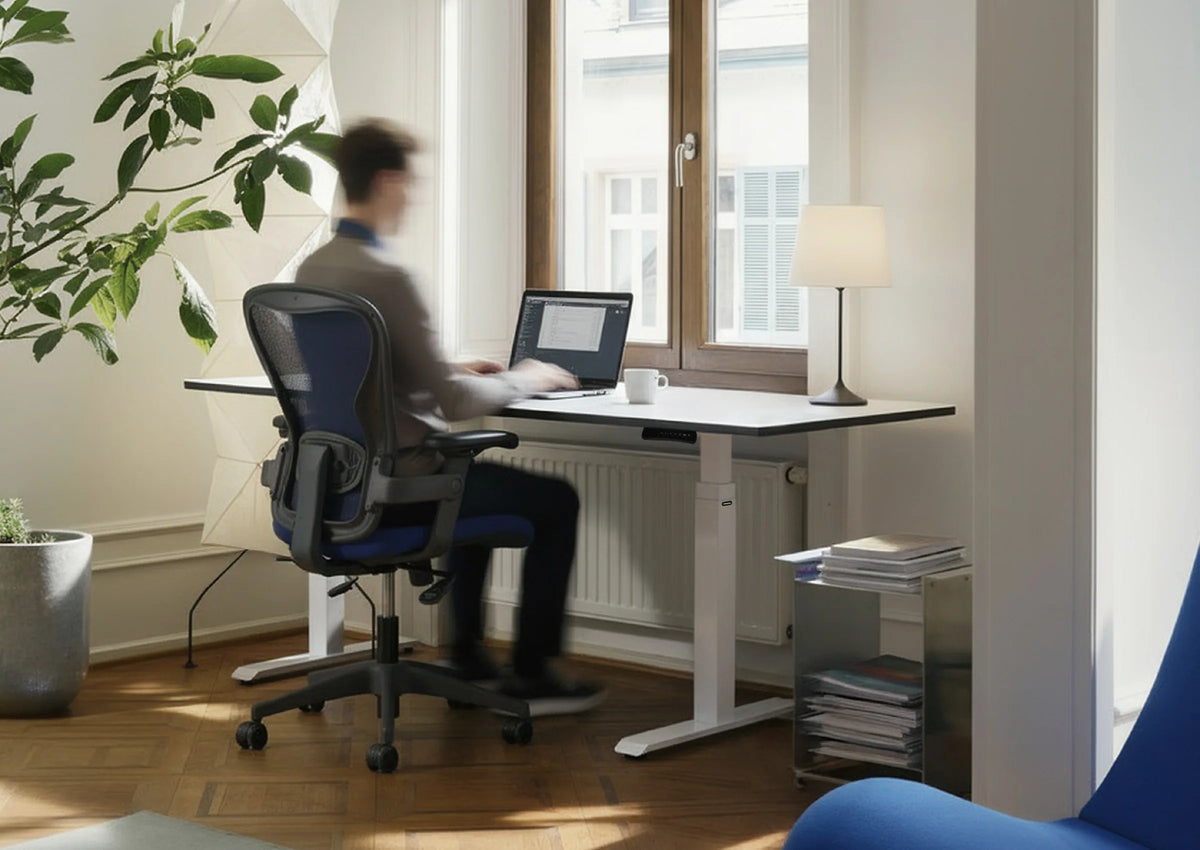 A person is seated at a modern, height-adjustable celexon E-Adjust 58123 desk, working on a laptop in a bright, tidy home office. Daylight streams in through a large window.