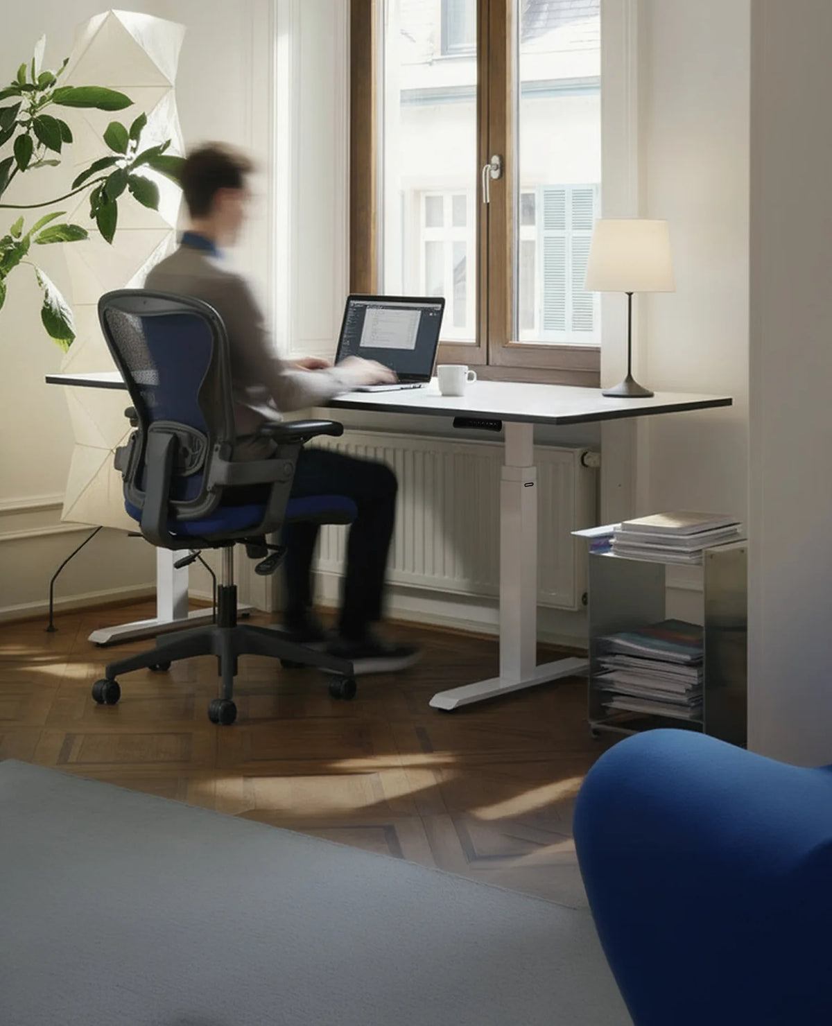 A person is seated at a modern, height-adjustable celexon E-Adjust 58123 desk, working on a laptop in a bright, tidy home office. Daylight streams in through a large window.