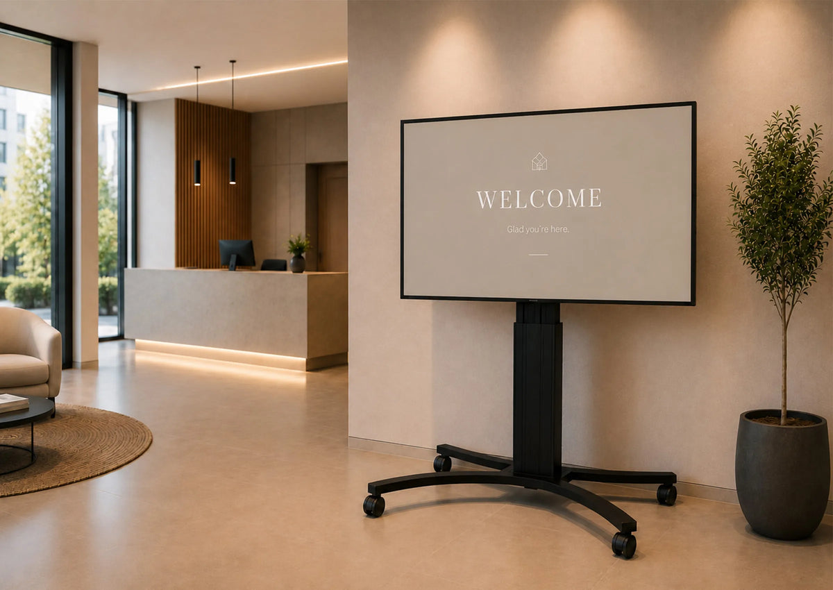 A premium reception area featuring a celexon Adjust-4286MB display stand shown from the front. The large display presents a clean, minimalist welcome screen. Light-colored materials, natural wood accents, elegant lighting, and a modern reception desk create a sophisticated, high-end atmosphere with a clear focus on the product.