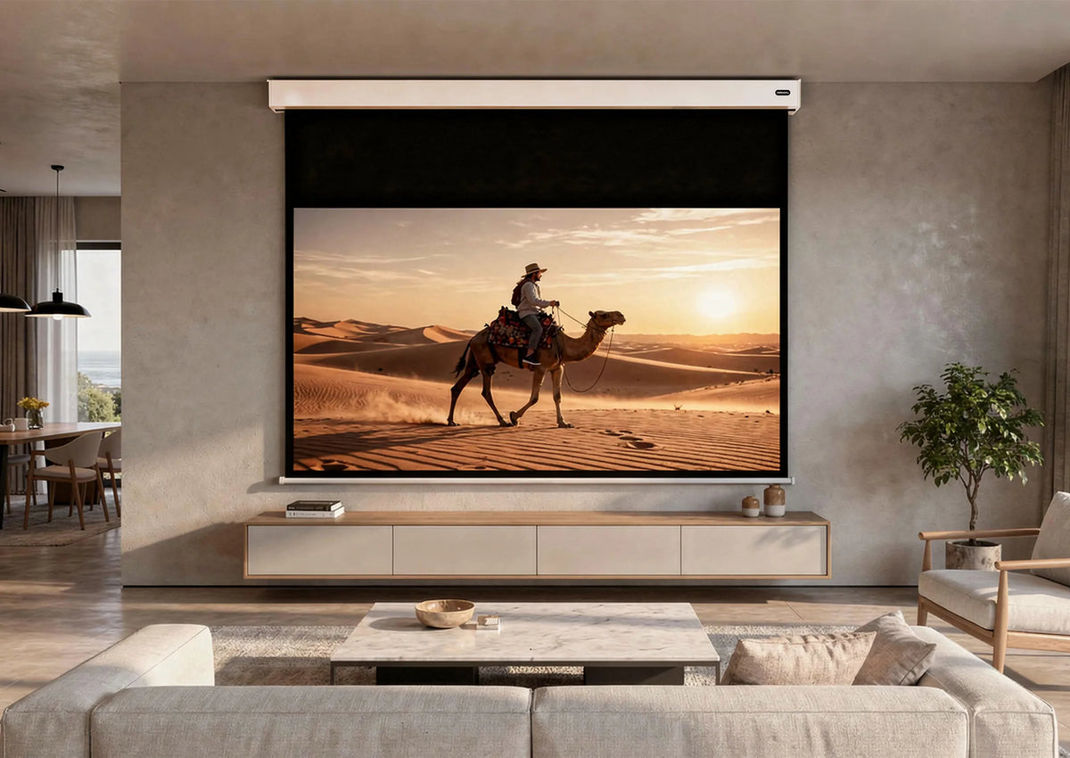 A modern living room with a celexon Electric Professional Plus V2.0 Battery projector screen. Displayed on it is a warm desert scene at sunset: a person riding a camel through sand dunes.