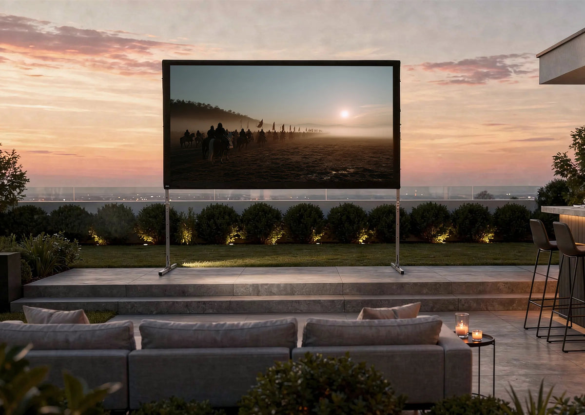 Luxurious outdoor home cinema in the evening: in a modernly designed garden, a celexon Folding Frame Projector Screen Mobile Expert with rear projection is set up on a terrace, displaying an atmospheric film scene featuring a rider at sunrise. In front of it, elegant outdoor sofas with cushions and candles invite relaxed viewing. On the right, a stylish illuminated outdoor bar with bar stools, glasses, and bottles complements the scene.
