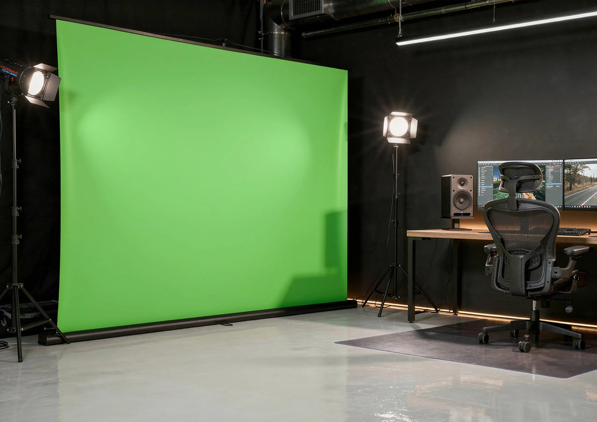 A professionally designed studio and work environment, with a large, free-standing celexon Mobile Lite Chroma Key green screen positioned on the left, while on the right there is a modern workstation featuring a minimalist wooden desk, two monitors displaying video editing software, and two speakers.