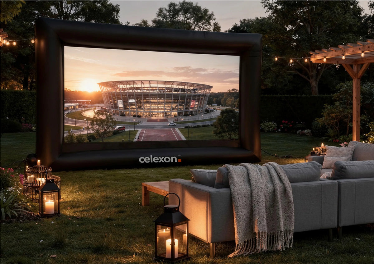 Atmospherically staged outdoor home cinema in a garden at dusk. At the center is the celexon inflatable outdoor projector screen INF200, displaying a modern stadium bathed in the warm light of the sunset. In front of it, cozy sofas with blankets invite people to sit, while lanterns with candles and string lights create a homely atmosphere.