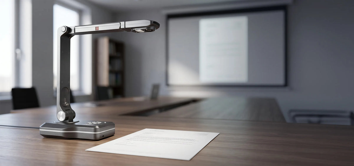 A modern celexon DK800 document camera on a conference table next to a document; in the background, a meeting room and projection screen are visible out of focus.