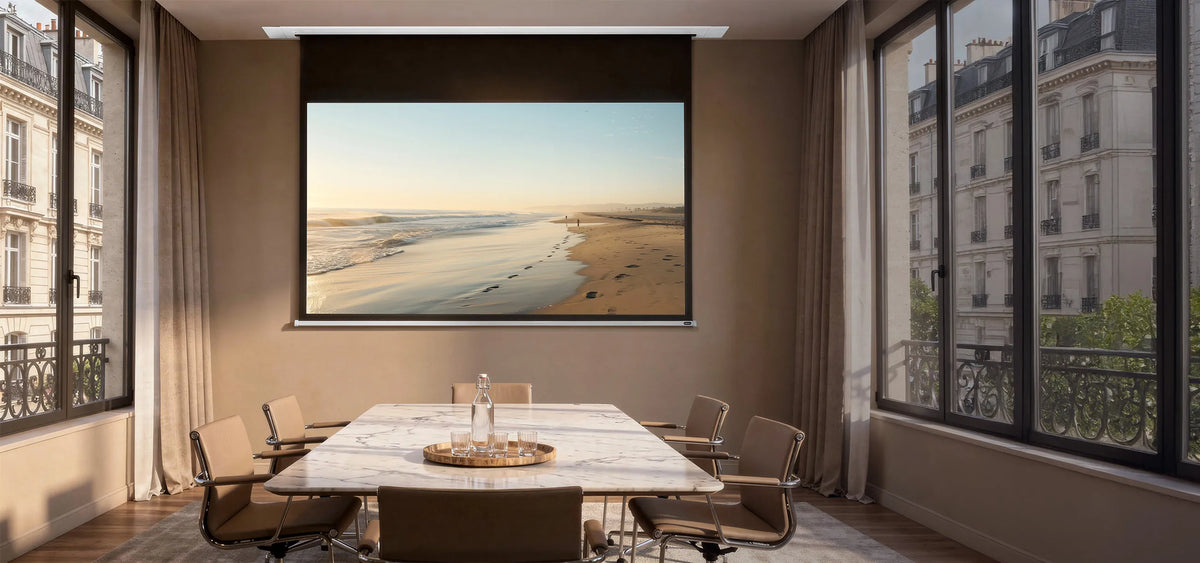 An elegant meeting room with floor-to-ceiling windows overlooking classic city façades. At the center stands a rectangular marble table surrounded by modern chairs. A celexon Ceiling Recessed Electric Professional Plus projector screen is integrated into the ceiling, displaying a calm beach scene with sea, sand, and footprints in warm morning or evening light.