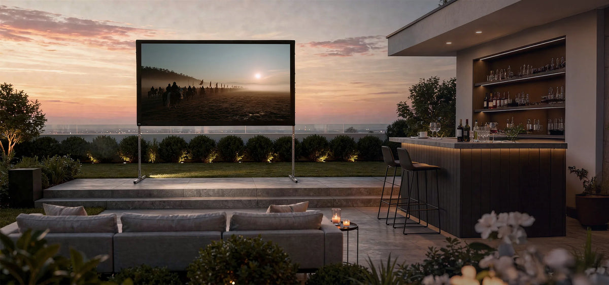 Luxurious outdoor home cinema in the evening: in a modernly designed garden, a celexon Folding Frame Projector Screen Mobile Expert with rear projection is set up on a terrace, displaying an atmospheric film scene featuring a rider at sunrise. In front of it, elegant outdoor sofas with cushions and candles invite relaxed viewing. On the right, a stylish illuminated outdoor bar with bar stools, glasses, and bottles complements the scene.
