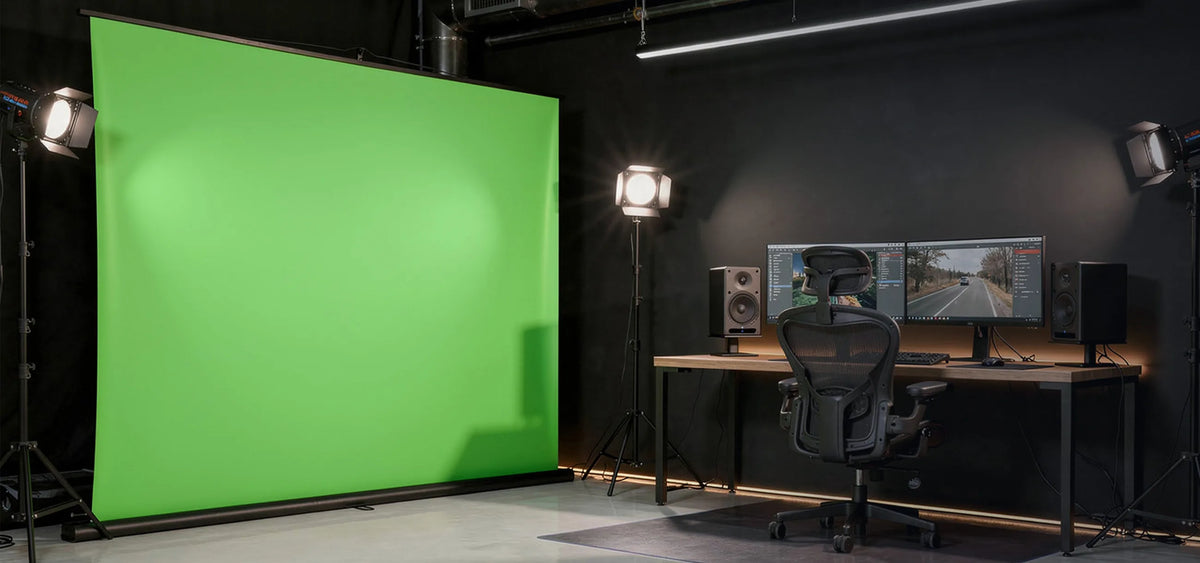 A professionally designed studio and work environment, with a large, free-standing celexon Mobile Lite Chroma Key green screen positioned on the left, while on the right there is a modern workstation featuring a minimalist wooden desk, two monitors displaying video editing software, and two speakers.