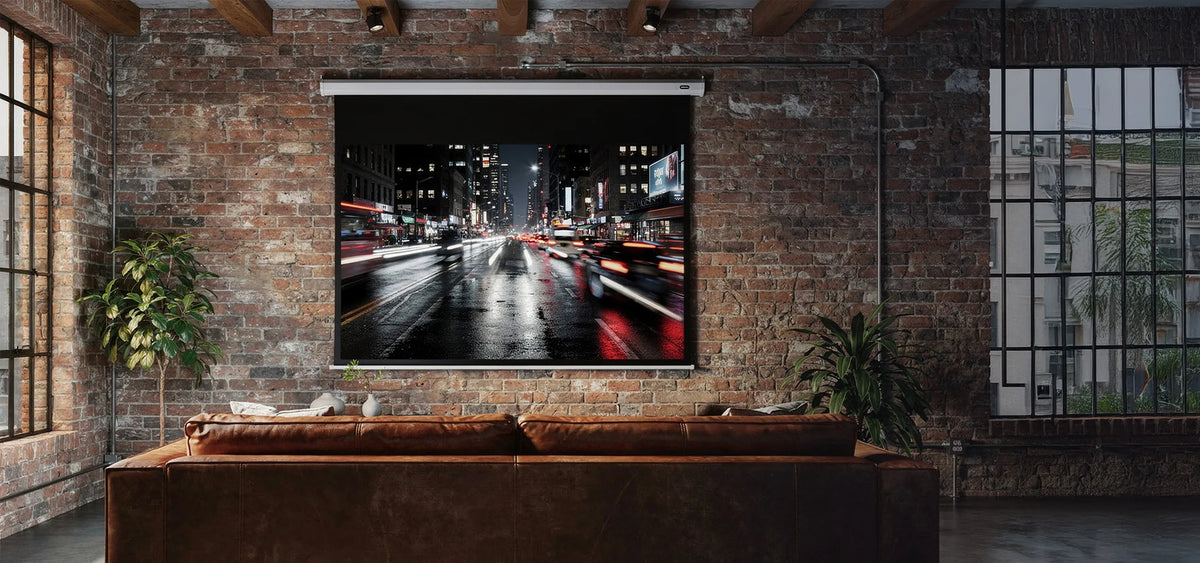The image shows an industrial-style living room with a brick wall, a leather sofa, and a large celexon Projector Screen Electric Economy, displaying a nighttime city street with traffic light trails.