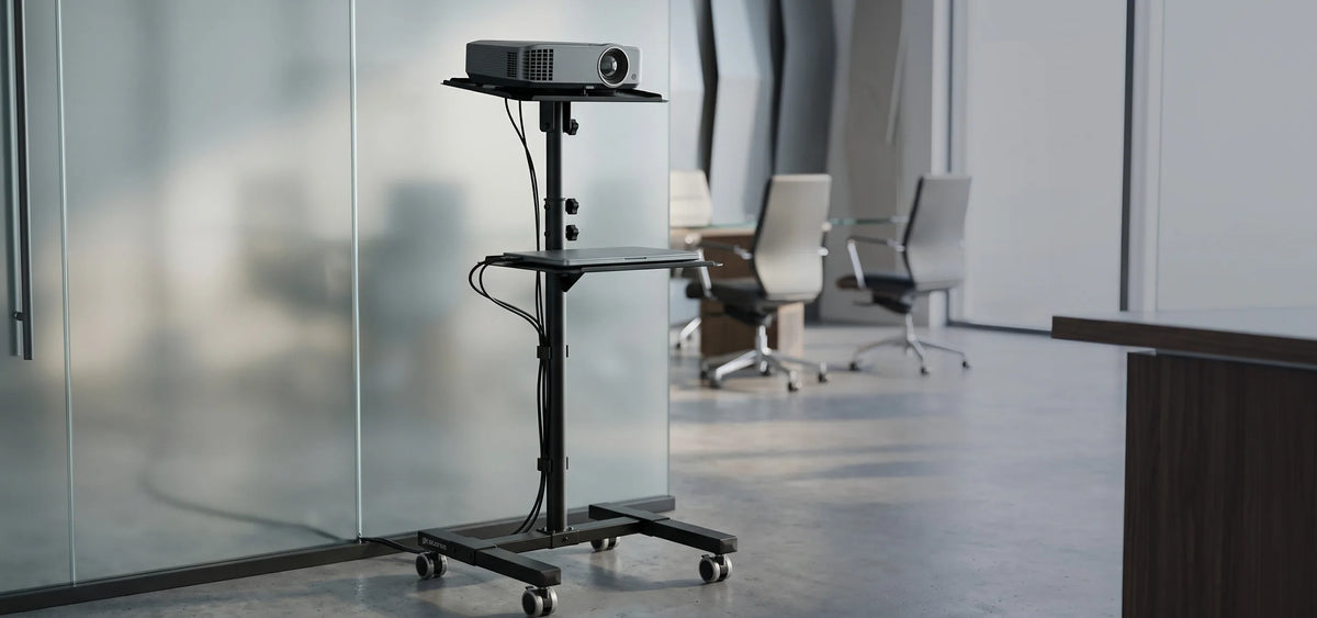 A mobile celexon PT3020 projector trolley with castors and height-adjustable shelves holding a projector and a laptop. The trolley is positioned in a modern office environment with glass walls and a meeting room in the background.