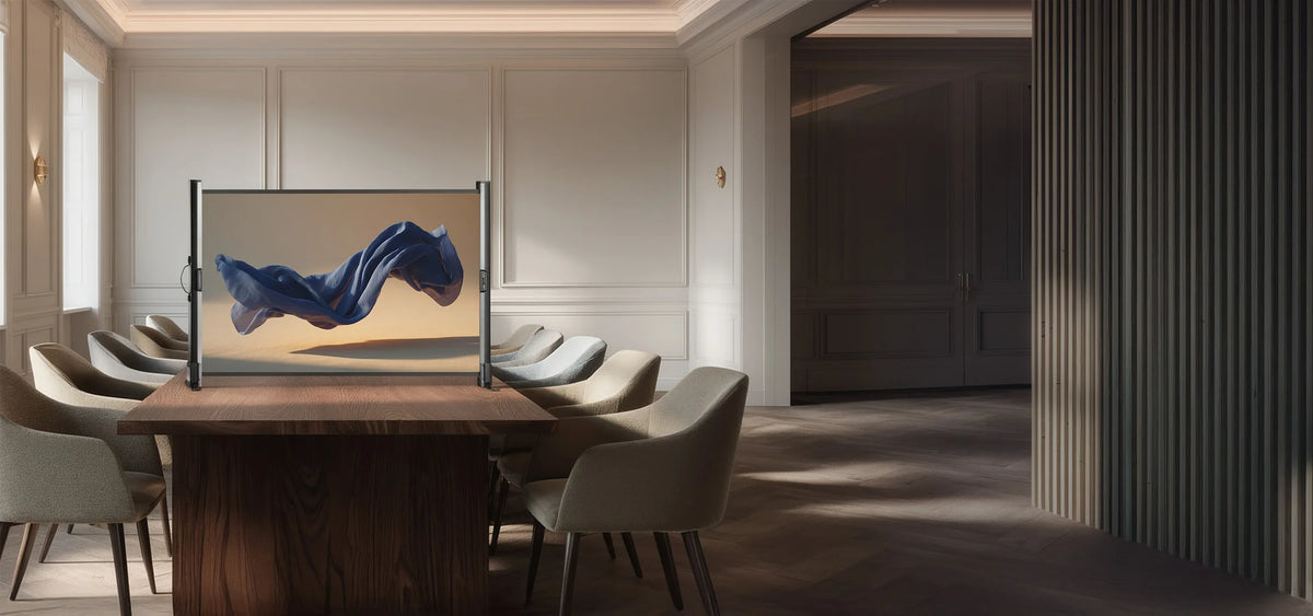 Classically modern meeting room with light-colored walls and fine stucco details. In the center of the room stands a large wooden table, on which a celexon Mobile Professional tabletop screen is placed. The projection shows a floating, softly flowing blue fabric.