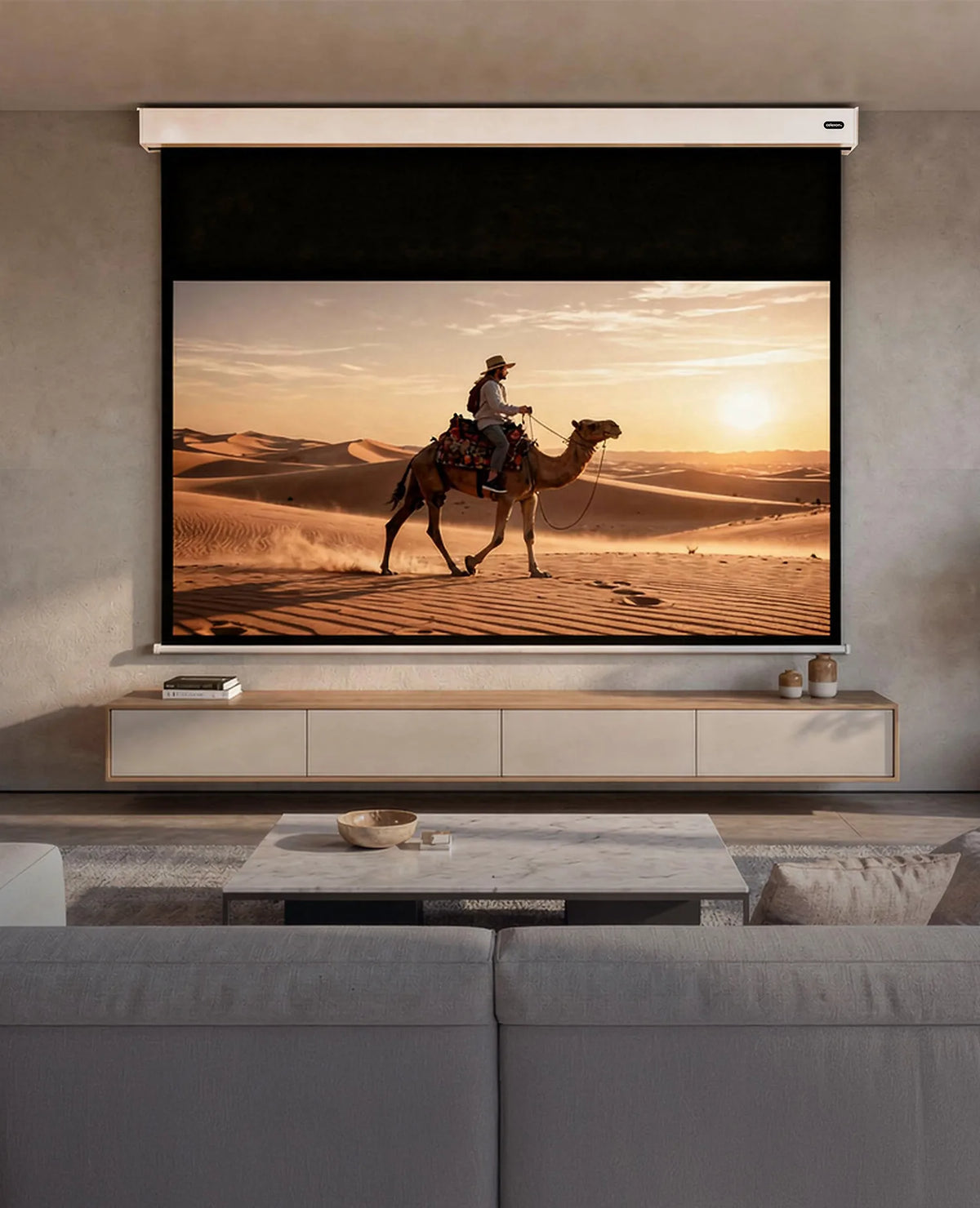 A modern living room with a celexon Electric Professional Plus V2.0 Battery projector screen. Displayed on it is a warm desert scene at sunset: a person riding a camel through sand dunes.