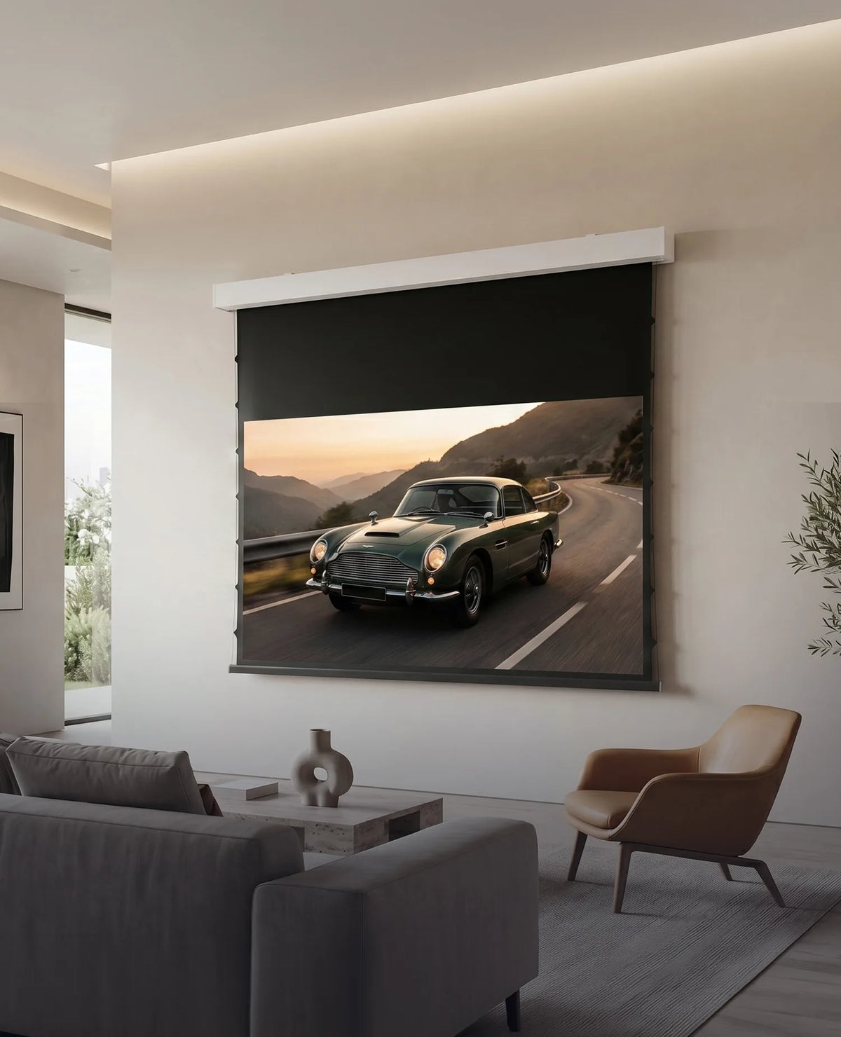 A modern living room featuring a celexon DARKVISION ALR Premium Plus Screen Electric Tension, displaying a classic sports car on a winding mountain road. Light-colored seating, a leather armchair, and a large indoor plant complement the elegant, minimalist interior.