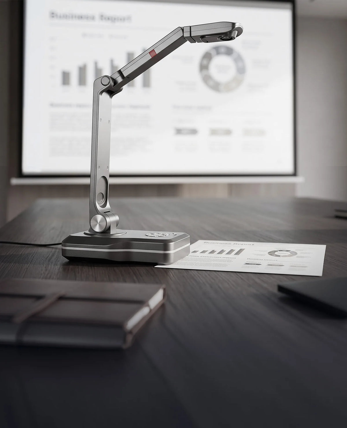A modern meeting room with a celexon DK500 document camera on a conference table; next to it is a business report whose contents are displayed on a large projection screen in the background.