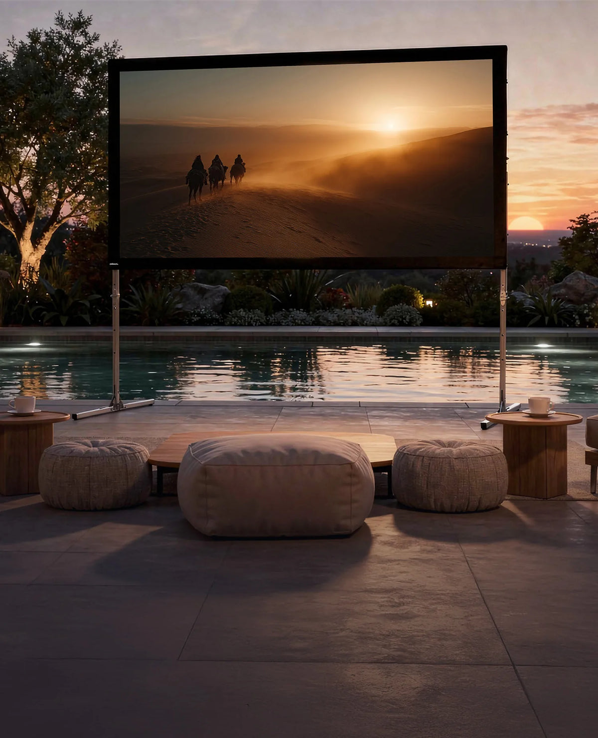 An exclusive outdoor home cinema at sunset: in front of a stylishly illuminated pool, a celexon Folding Frame Screen Mobile Expert set up for front projection is installed, displaying an atmospheric film scene with riders in a desert landscape bathed in warm evening light. In front of it are low seating arrangements and small side tables with drinks.