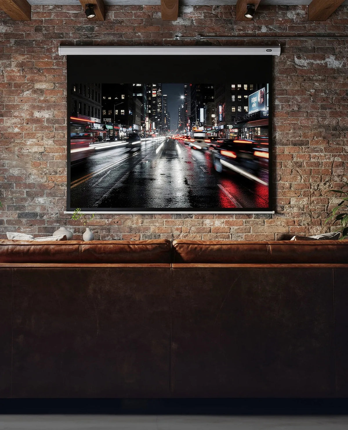 The image shows an industrial-style living room with a brick wall, a leather sofa, and a large celexon Projector Screen Electric Economy, displaying a nighttime city street with traffic light trails.