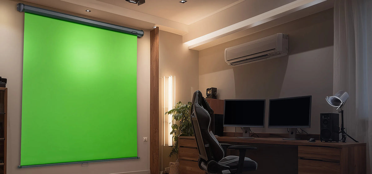 The image shows a modern studio workspace in which, on the left, a large, wall-mounted celexon manual Chroma Key green screen is installed, while on the right a tidy workstation with a wooden desk, two monitors, an office chair, a plant, and an air-conditioning unit creates a professional environment for video and content production.