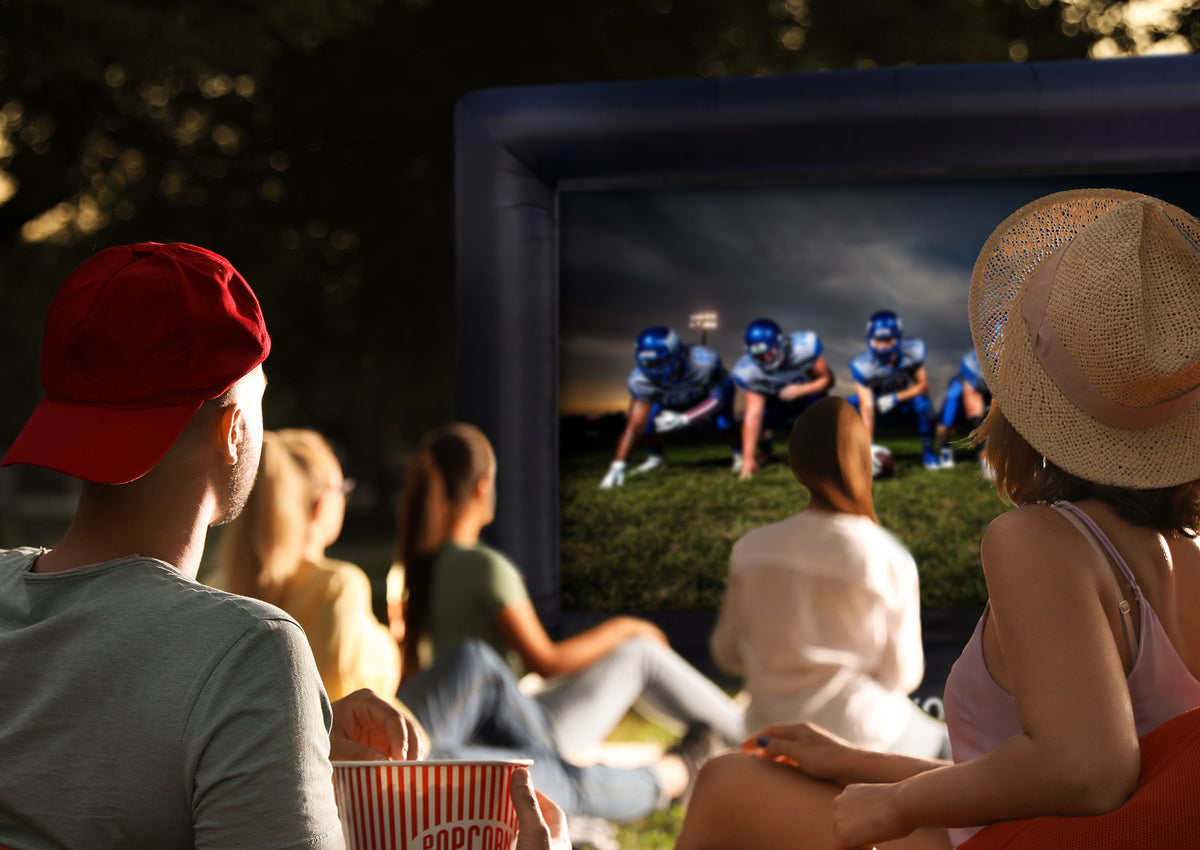Atmospheric outdoor public viewing scene at dusk: a group of people sits relaxed on the grass, watching a celexon inflatable outdoor projector screen INF200 that displays an American football game with players in a starting formation under floodlights.