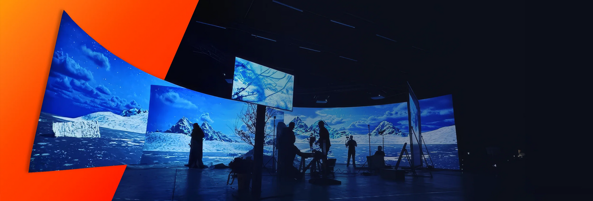 A large, dark studio with multiple curved projection screens displaying an icy polar landscape with mountains and sea; in front of them stand several silhouetted people and film equipment.