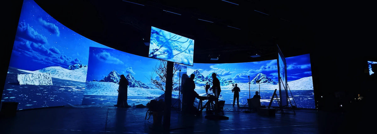 A large, dark studio with multiple curved projection screens displaying an icy polar landscape with mountains and sea; in front of them stand several silhouetted people and film equipment.