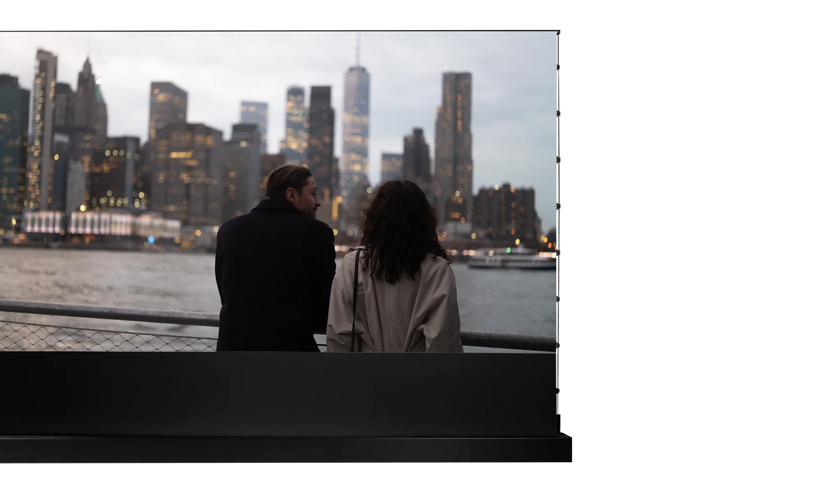 Extended celexon HomeCinema Plus floor-rising scissor screen. The projection shows two people standing with their backs to the camera at a railing by the water, looking out at a skyline of tall, closely spaced high-rise buildings.