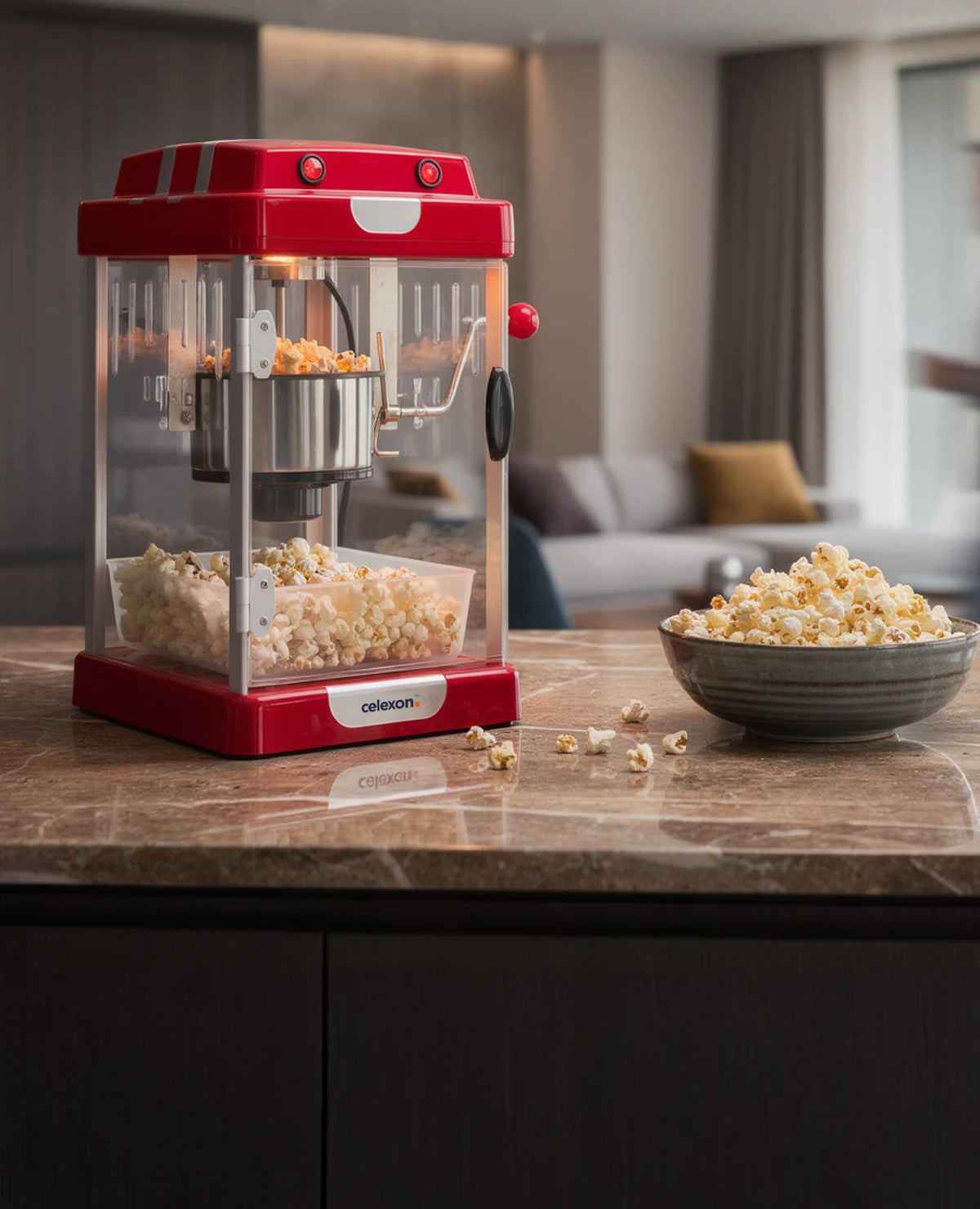 In the foreground, a red celexon CP1000 popcorn machine stands on a dark stone countertop, next to a bowl of freshly popped popcorn, casually and invitingly arranged. The modern living space in the background is deliberately kept out of focus.