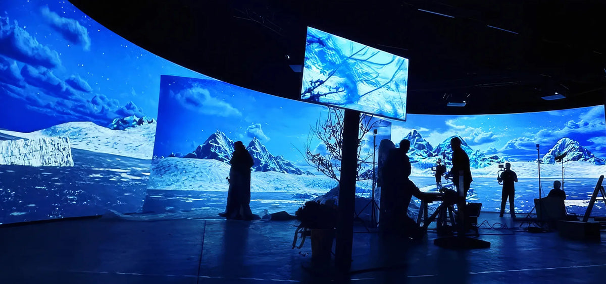 A large, dark studio with multiple curved projection screens displaying an icy polar landscape with mountains and sea; in front of them stand several silhouetted people and film equipment.