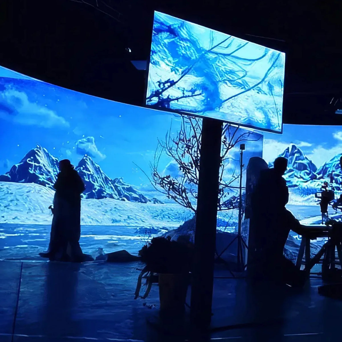 A large, dark studio with multiple curved projection screens displaying an icy polar landscape with mountains and sea; in front of them stand several silhouetted people and film equipment.