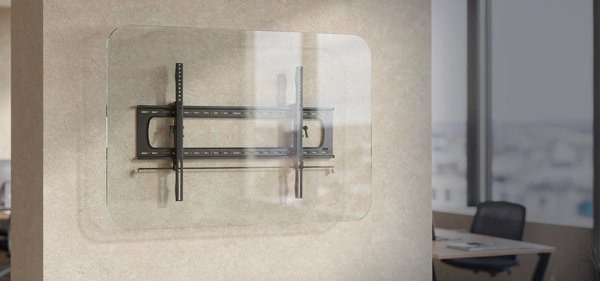The image shows the celexon TV/display wall mount Adjust/Fixed attached to a transparent glass panel. The bracket consists of a sturdy metal construction with horizontal and vertical rails for mounting a screen. The scene is set in a modern office or workspace with a neutral wall, large windows, and a desk in the background.