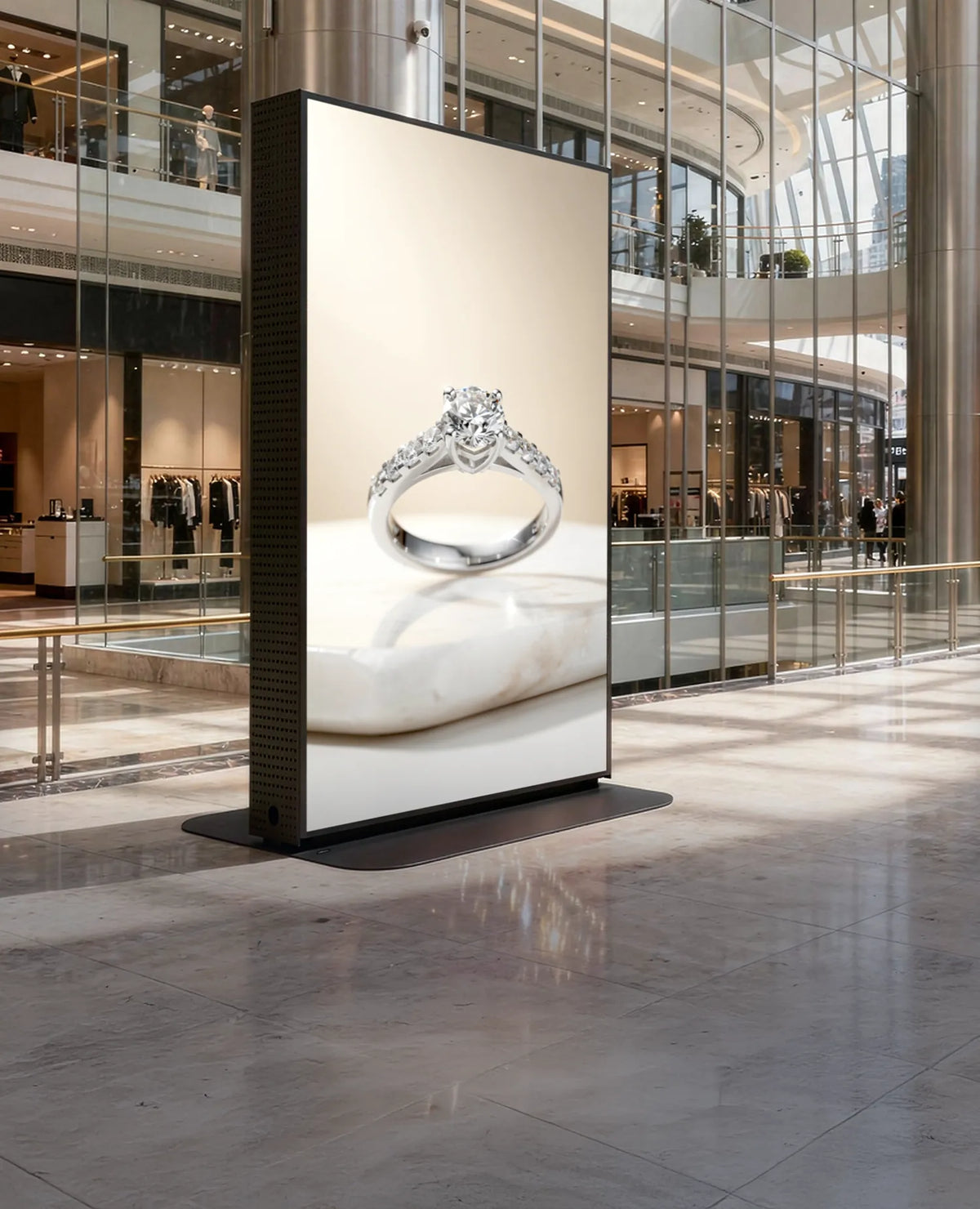 The image shows a large, freestanding digital advertising display in a modern shopping mall with high glass architecture and open galleries. On the vertical screen, an elegant advertisement of a diamond ring is presented. Boutiques and shop windows surround the display.