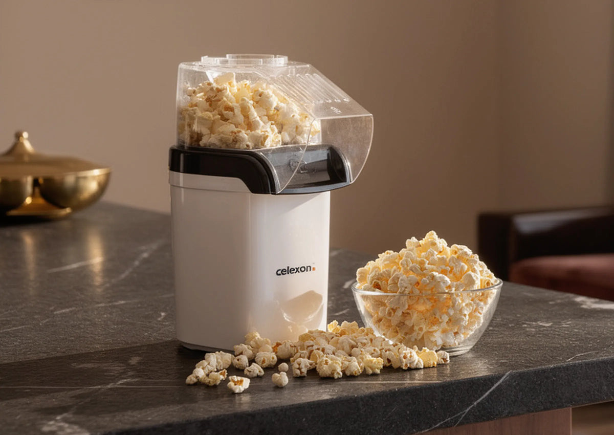 In the foreground, a compact celexon CP150 popcorn machine stands on a dark kitchen or bar counter. Freshly popped popcorn fills the transparent container, with a glass bowl of popcorn beside it and a few kernels casually scattered on the surface. In the background, a cozy living space with comfortable armchairs and a floor lamp can be seen, softly out of focus.