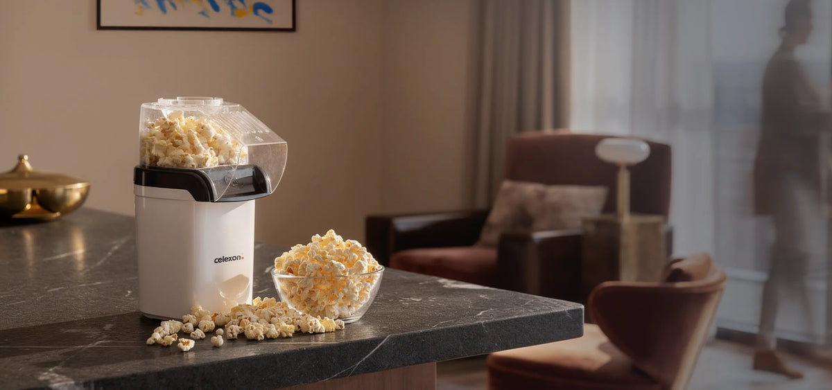 In the foreground, a compact celexon CP150 popcorn machine stands on a dark kitchen or bar counter. Freshly popped popcorn fills the transparent container, with a glass bowl of popcorn beside it and a few kernels casually scattered on the surface. In the background, a cozy living space with comfortable armchairs and a floor lamp can be seen, softly out of focus.