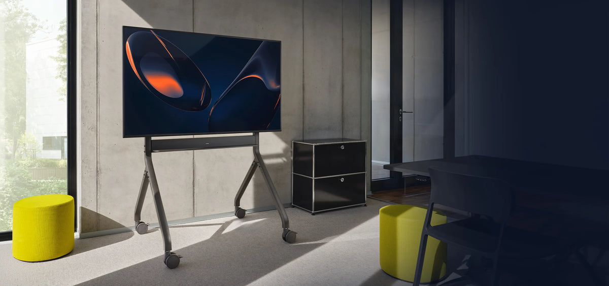 The image shows a modern, mobile display mounted on a celexon Expert electrically height-adjustable display trolley Oslo in a bright, minimalist room. On the screen, an abstract, color-rich visual is displayed, while the surroundings feature a concrete wall, large windows, and colorful seating stools.