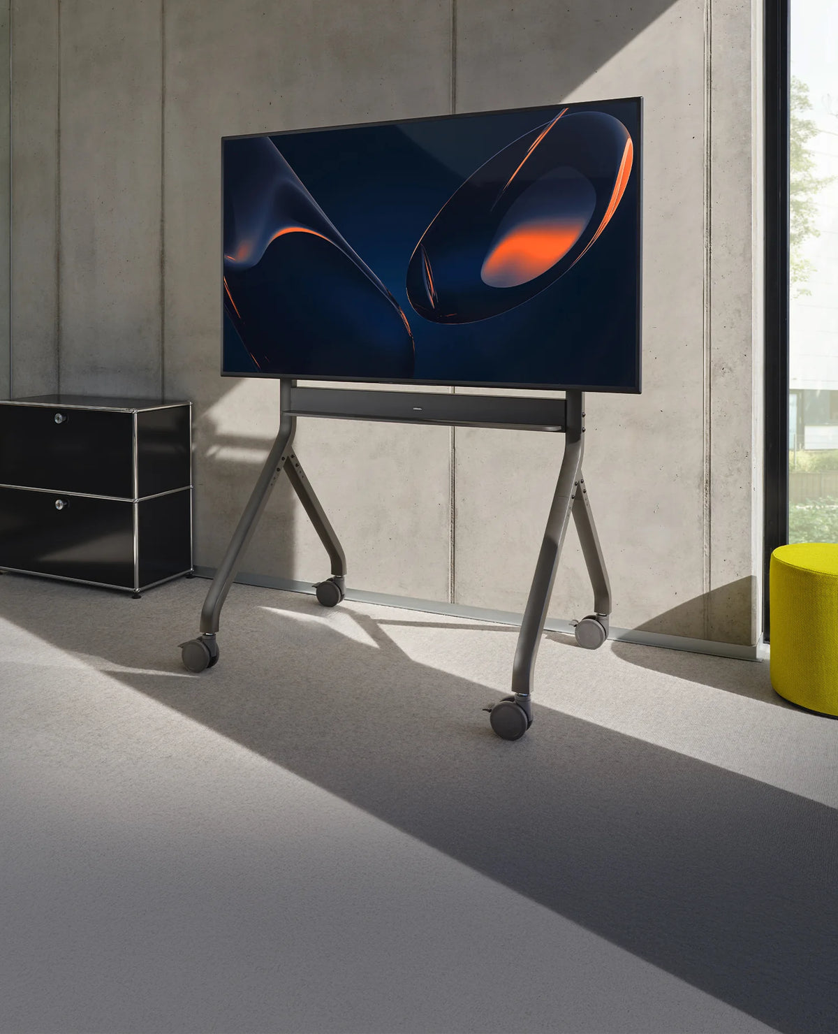 The image shows a modern, mobile display mounted on a celexon Expert electrically height-adjustable display trolley Oslo in a bright, minimalist room. On the screen, an abstract, color-rich visual is displayed, while the surroundings feature a concrete wall, large windows, and colorful seating stools.