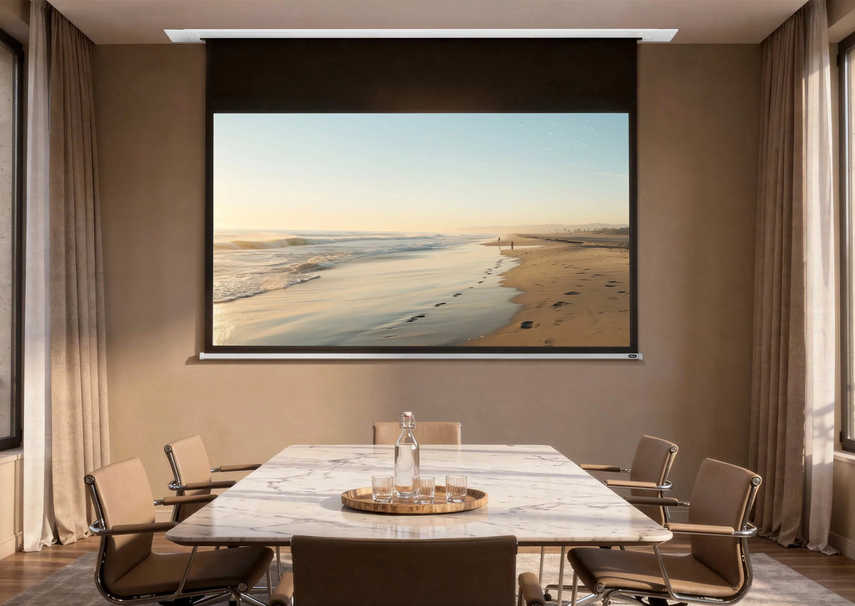 An elegant meeting room with floor-to-ceiling windows overlooking classic city façades. At the center stands a rectangular marble table surrounded by modern chairs. A celexon Ceiling Recessed Electric Professional Plus projector screen is integrated into the ceiling, displaying a calm beach scene with sea, sand, and footprints in warm morning or evening light.