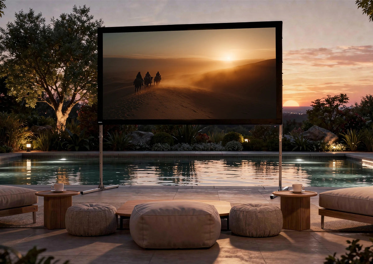 An exclusive outdoor home cinema at sunset: in front of a stylishly illuminated pool, a celexon Folding Frame Screen Mobile Expert set up for front projection is installed, displaying an atmospheric film scene with riders in a desert landscape bathed in warm evening light. In front of it are low seating arrangements and small side tables with drinks.