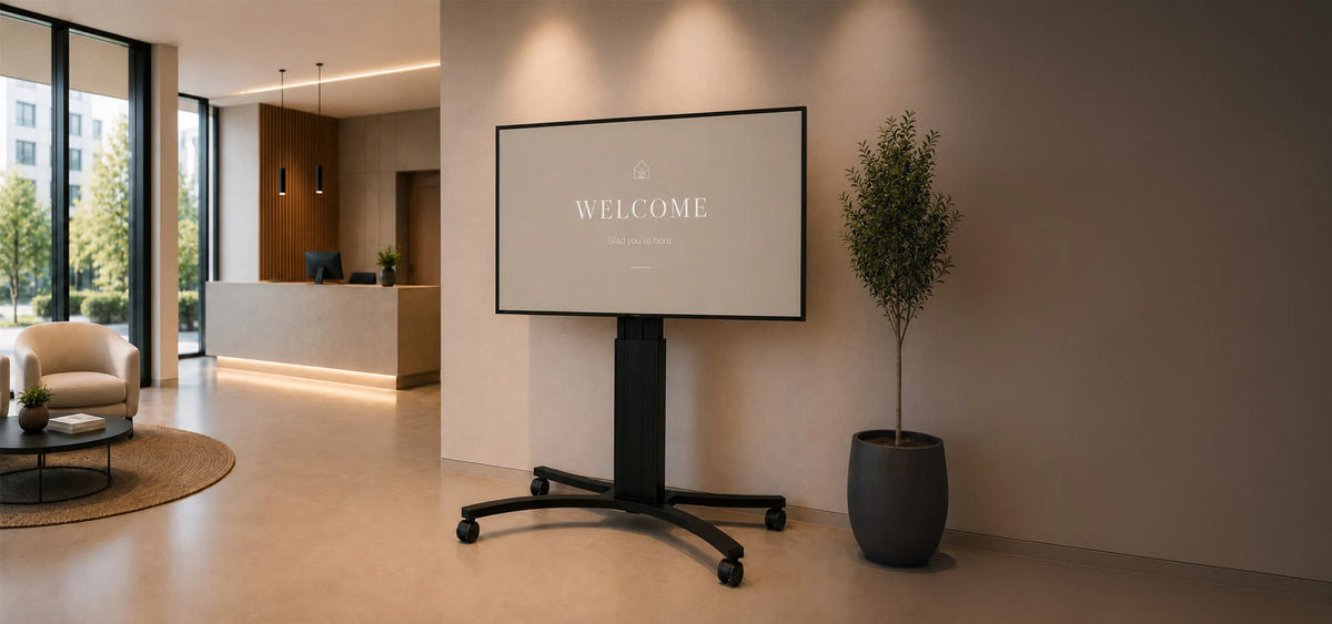 A premium reception area featuring a celexon Adjust-4286MB display stand shown from the front. The large display presents a clean, minimalist welcome screen. Light-colored materials, natural wood accents, elegant lighting, and a modern reception desk create a sophisticated, high-end atmosphere with a clear focus on the product.
