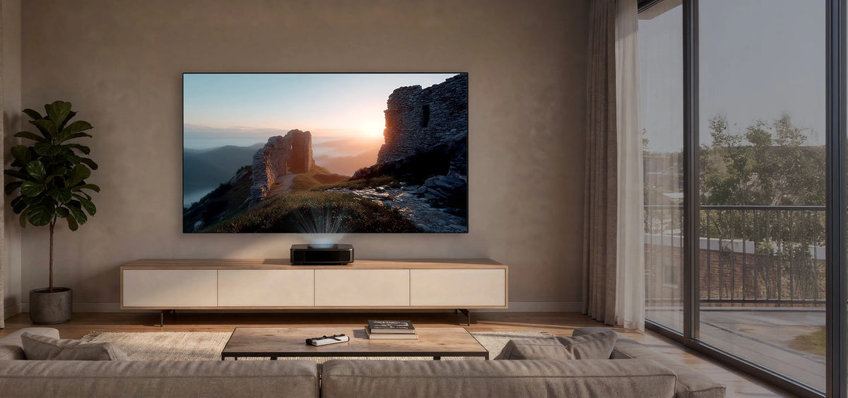 A modern, calmly designed living space. Mounted frontally on the wall is a celexon CLR HomeCinema Performance UST high-contrast fixed-frame projector screen. Displayed on it is a moody landscape: an ancient stone ruin complex on a hill, illuminated by the low sun. Below the projection stands a long, low sideboard in light wood with white fronts. Centered on it is a compact UST projector, projecting the image upward onto the screen.
