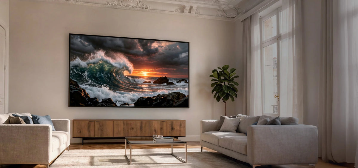 The image shows an elegant living room with two sofas and a celexon HomeCinema High Contrast Frame Projector Screen – Dynamic Slate ALR, displaying a powerful ocean wave at sunset.
