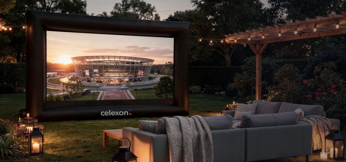 Atmospherically staged outdoor home cinema in a garden at dusk. At the center is the celexon inflatable outdoor projector screen INF200, displaying a modern stadium bathed in the warm light of the sunset. In front of it, cozy sofas with blankets invite people to sit, while lanterns with candles and string lights create a homely atmosphere.