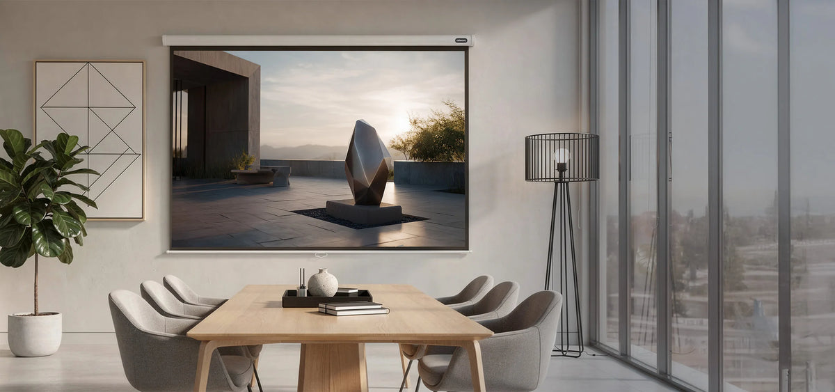 The image shows a modern meeting room with a wooden table and upholstered chairs, in which a celexon Manual Economy projector screen displays a calm outdoor architectural scene featuring an abstract sculpture in warm evening light.