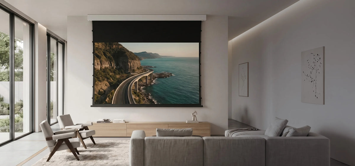 A modern living room featuring a celexon SOUNDVISION Premium Plus Screen Electric Tension, displaying a coastal road along the sea. Light-colored seating, a low sideboard, and minimalist furnishings create an elegant home cinema atmosphere.