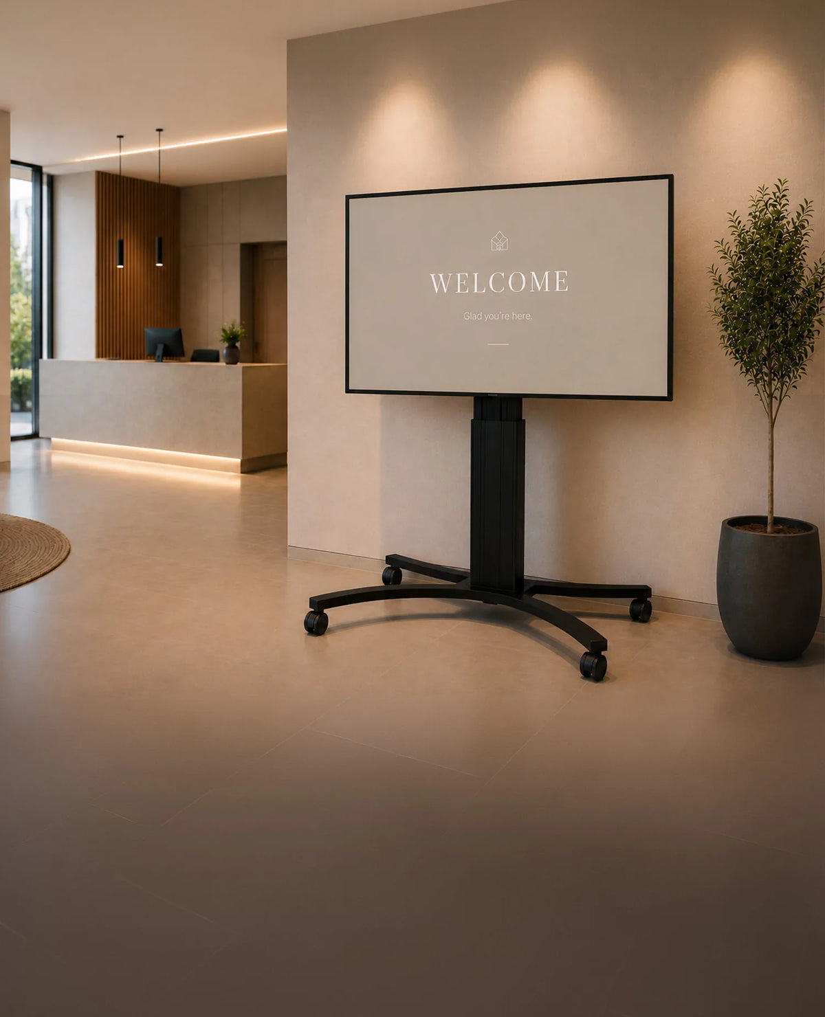 A premium reception area featuring a celexon Adjust-4286MB display stand shown from the front. The large display presents a clean, minimalist welcome screen. Light-colored materials, natural wood accents, elegant lighting, and a modern reception desk create a sophisticated, high-end atmosphere with a clear focus on the product.