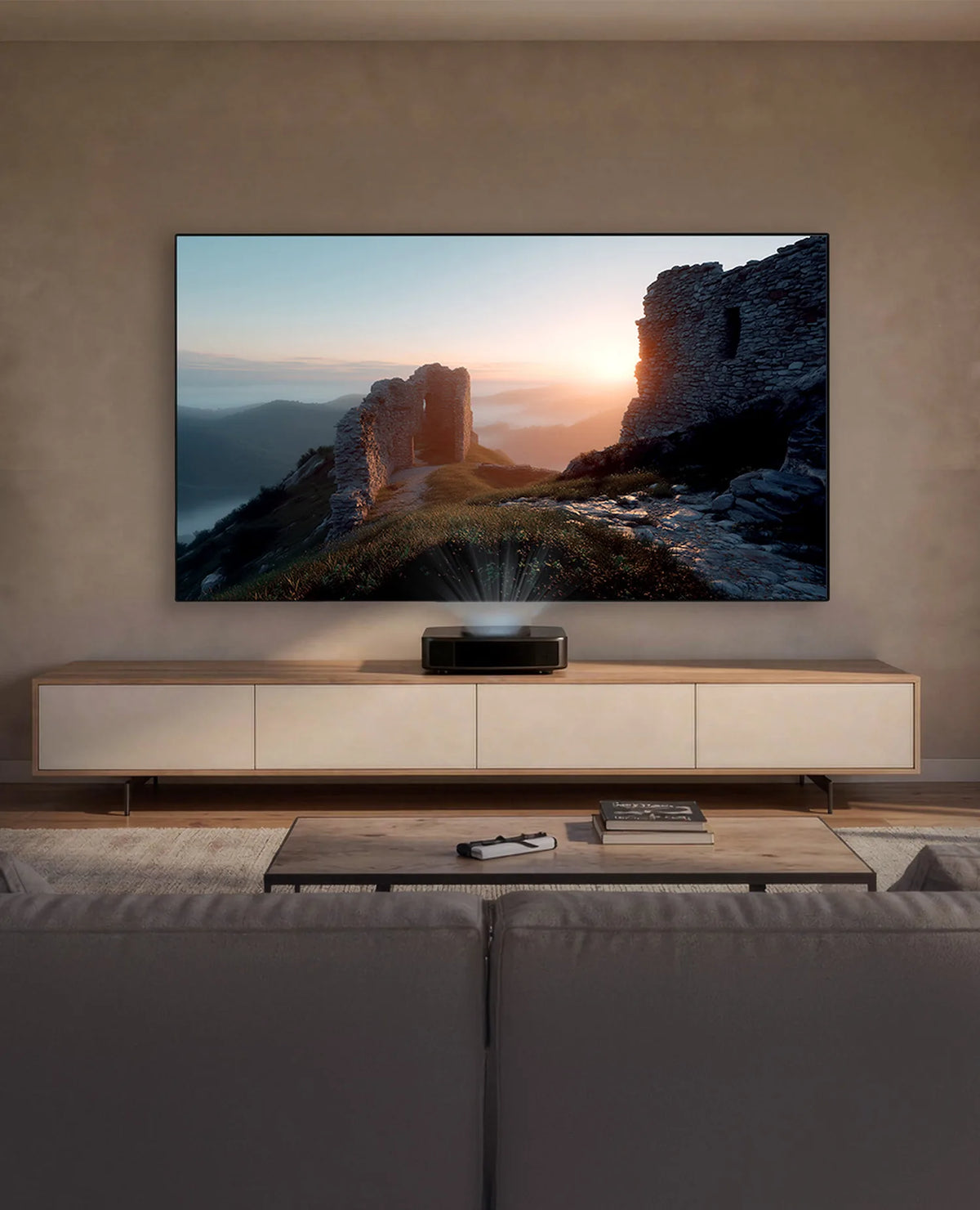 A modern, calmly designed living space. Mounted frontally on the wall is a celexon CLR HomeCinema Performance UST high-contrast fixed-frame projector screen. Displayed on it is a moody landscape: an ancient stone ruin complex on a hill, illuminated by the low sun. Below the projection stands a long, low sideboard in light wood with white fronts. Centered on it is a compact UST projector, projecting the image upward onto the screen.