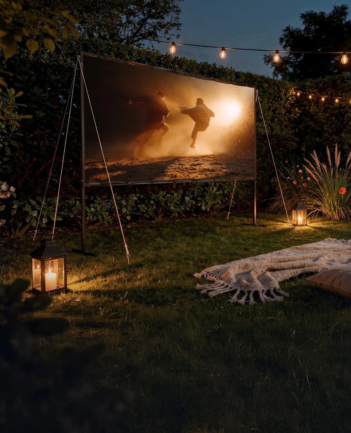 Atmospheric outdoor cinema in a garden at night: a celexon Basic Line folding frame screen is set up between plants and hedges, displaying a dynamic film scene with two people in action-packed motion against dusty, golden backlight. Warm string lights and lanterns illuminate the surroundings, while blankets, cushions, and rugs are laid out on the lawn.